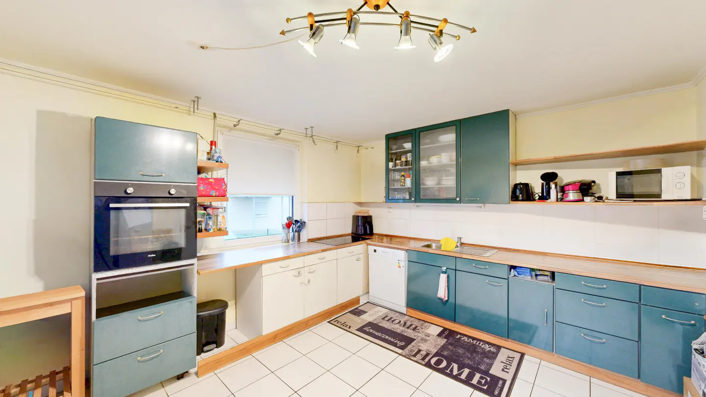 A bright kitchen with white tile floors, teal cabinets, and a "Home" rug. A window provides natural light.