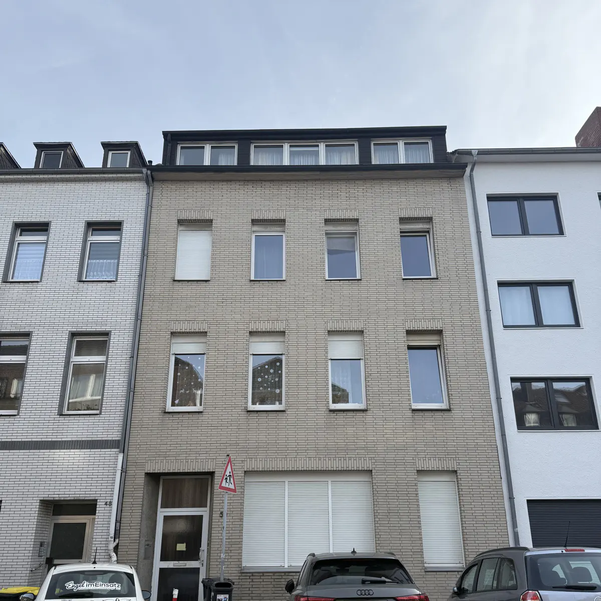 Three-story brick apartment building with a black roof and white window trim. Cars are parked in front.