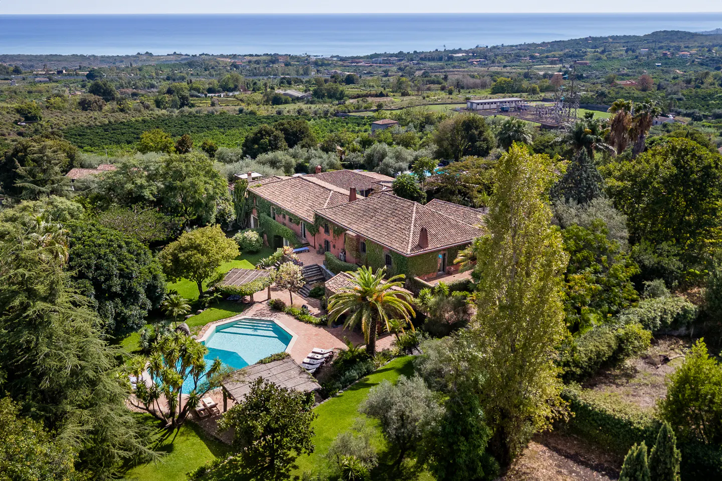 Aerial view of a terracotta villa with a pool, lush gardens, and palm trees, overlooking a coastal landscape and the sea.