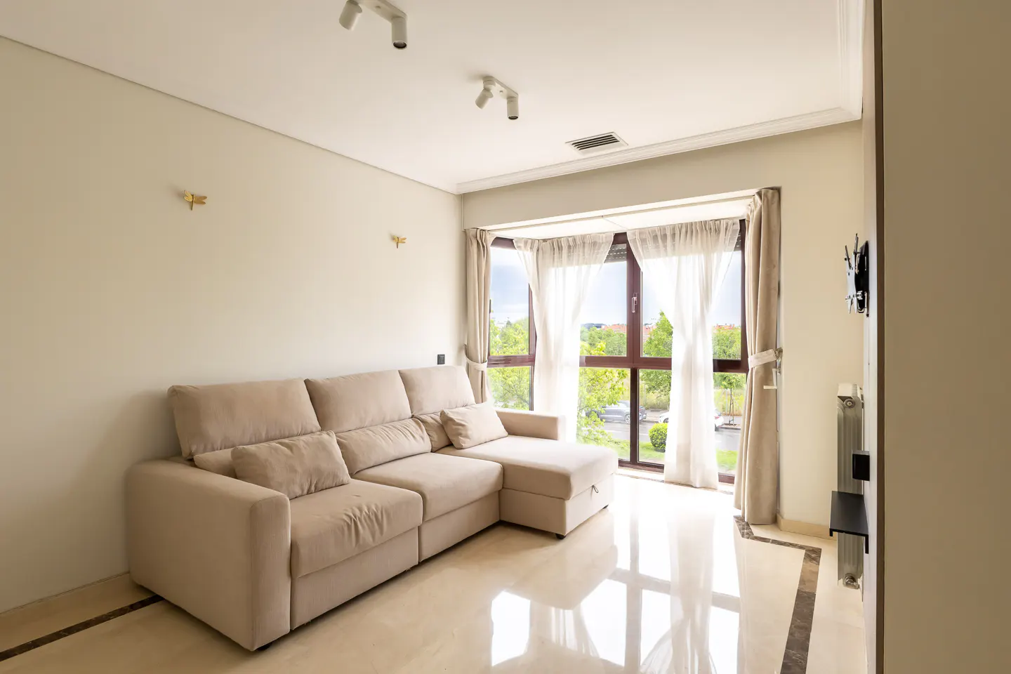 Living room with beige sectional sofa, marble floors, and large window with sheer curtains overlooking green trees.
