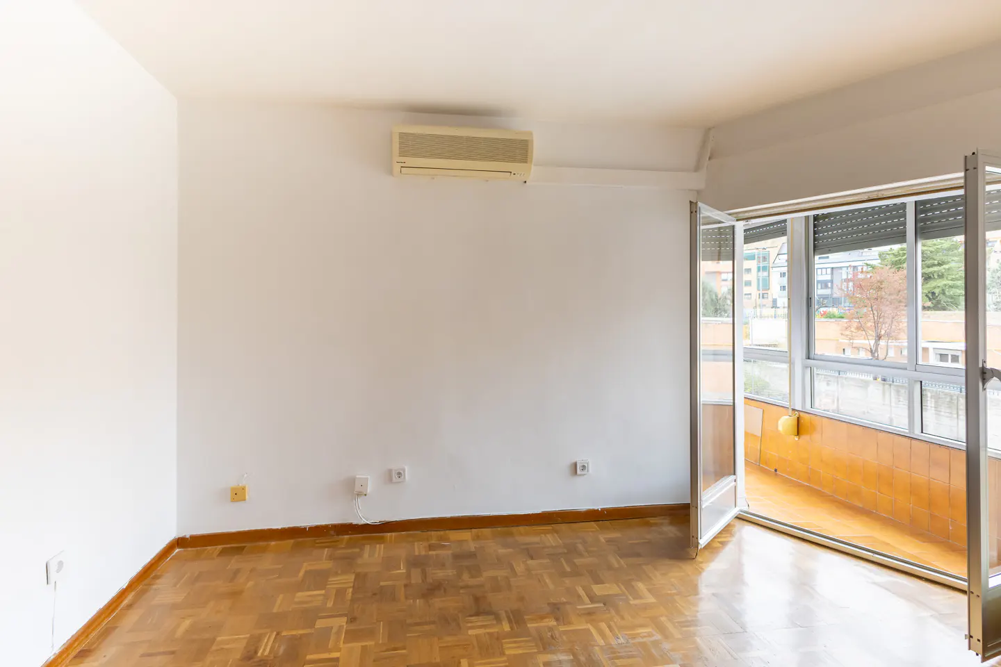 Bright, empty room with parquet floor, white walls, and open balcony doors. An air conditioner is mounted high on the wall.