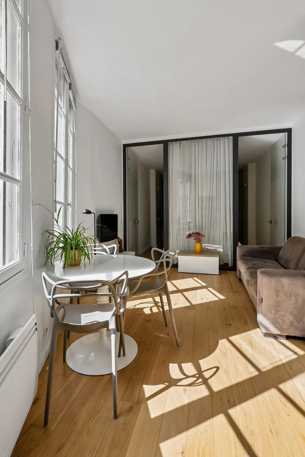 Bright, modern apartment interior with a round white table, chairs, and a brown sofa on a wood floor with sunlight streaming through windows.