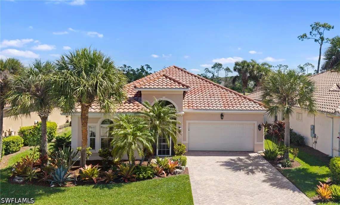 Beige single-story house with a red tile roof, white garage door, palm trees, and a brick driveway under a blue sky.