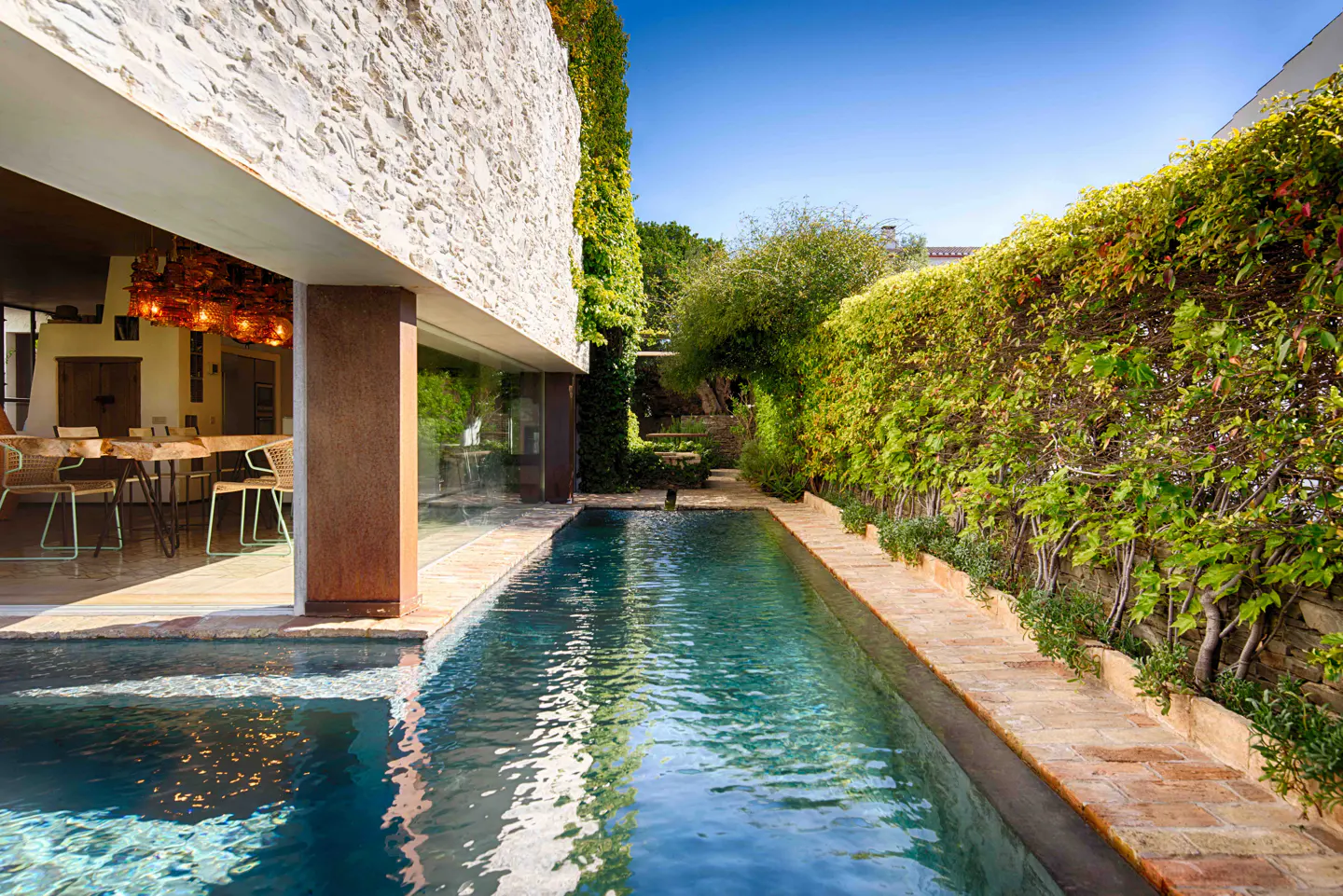 Outdoor pool with stone walls and green hedges. Dining table and chairs are visible through the open wall.