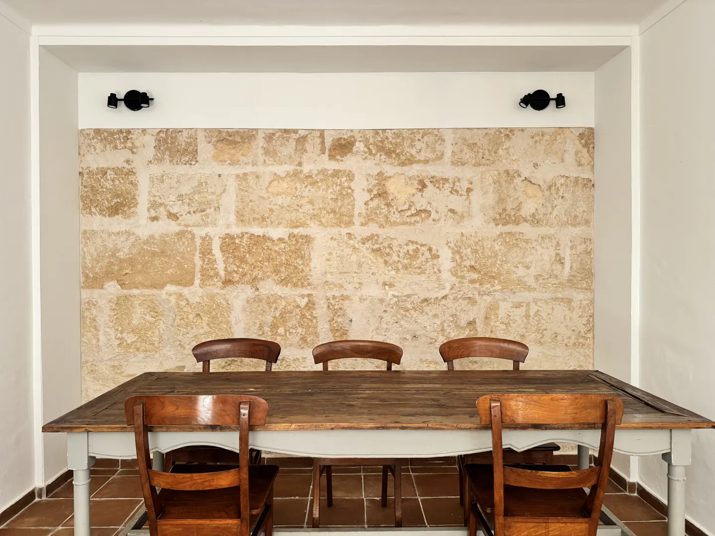 A dining room with a wooden table and chairs against a stone wall. Two black sconces are mounted above.