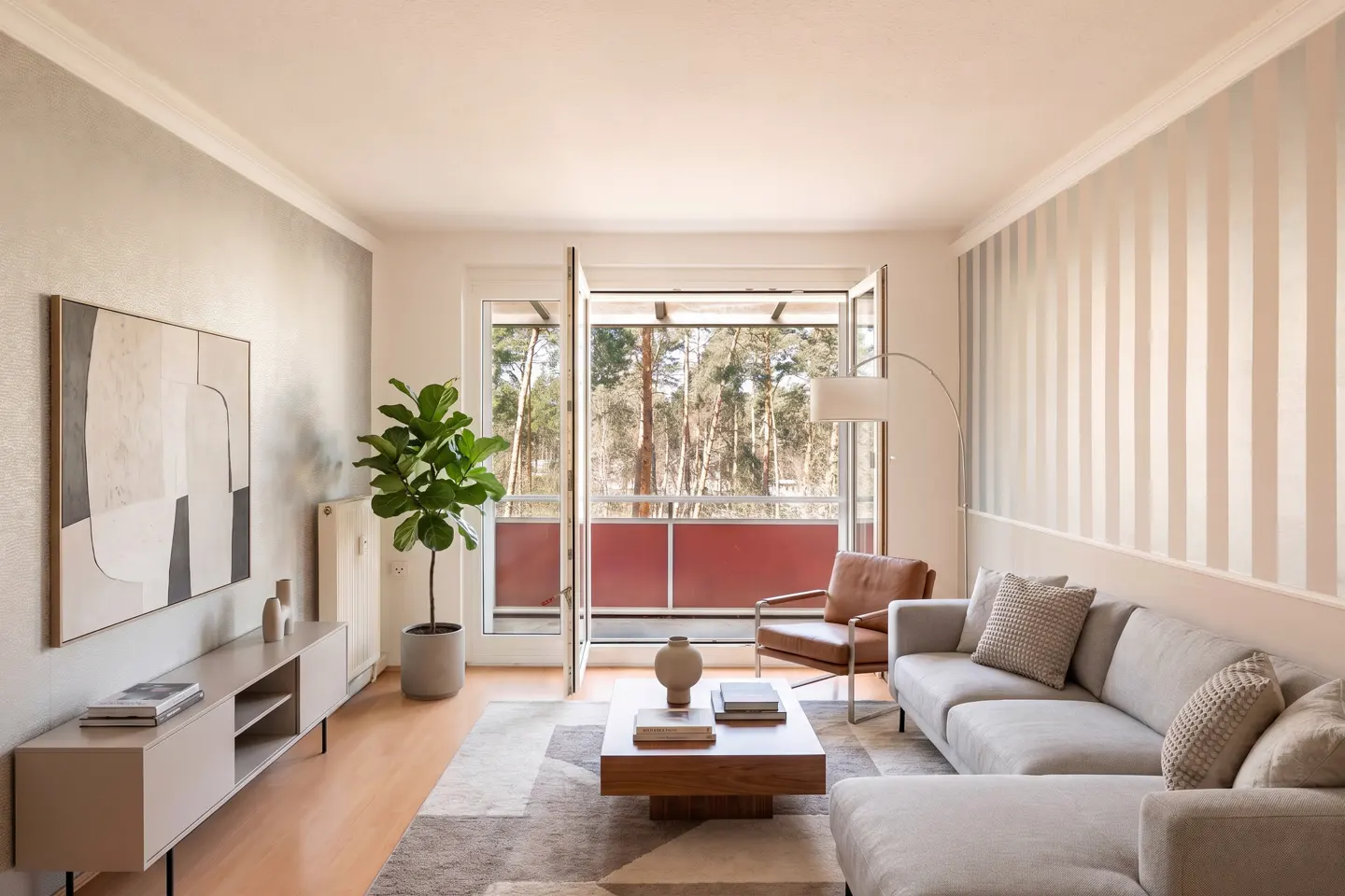 Bright living room with a gray sectional, brown leather chair, and a wooden coffee table. Balcony doors open to a forest view. A large abstract painting hangs on the wall.