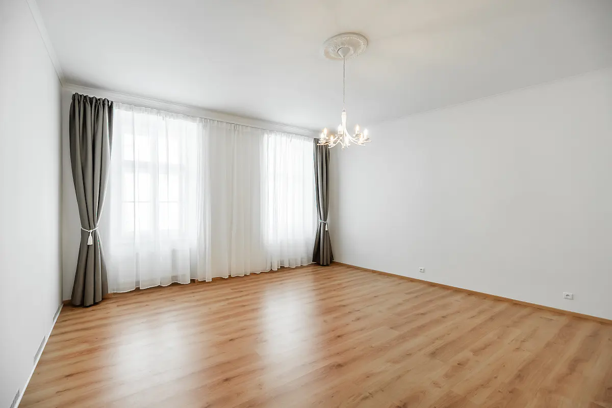 Bright, empty room with wood floors, white walls, and a chandelier. Windows have sheer white curtains and gray drapes.