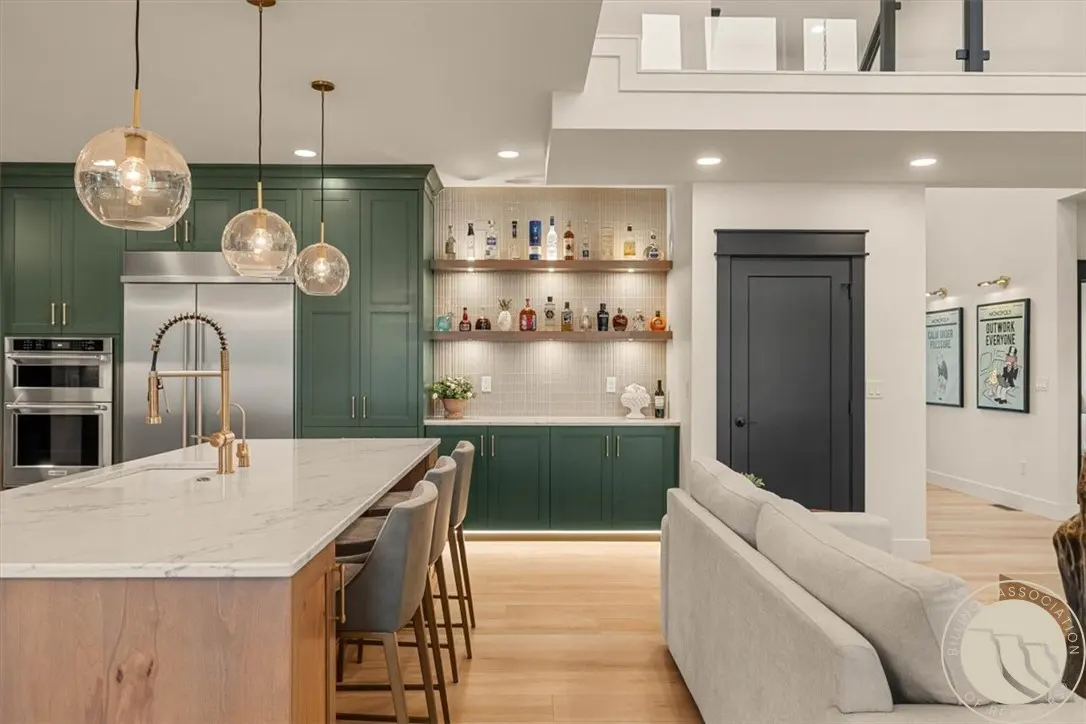 A modern kitchen with green cabinets, a marble island, and pendant lights. Shelves display liquor bottles. A gray sofa sits nearby.