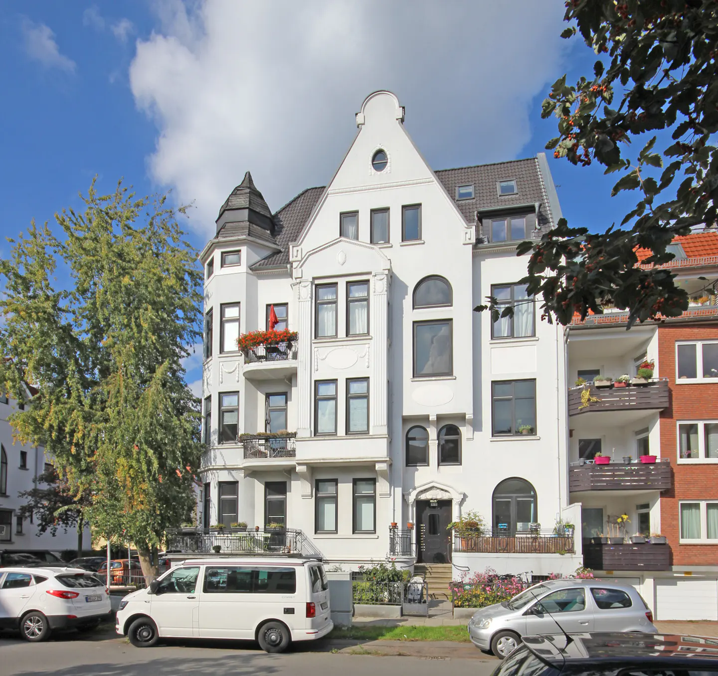 A three-story white apartment building with black trim and a turret on the left side. Cars are parked in front.