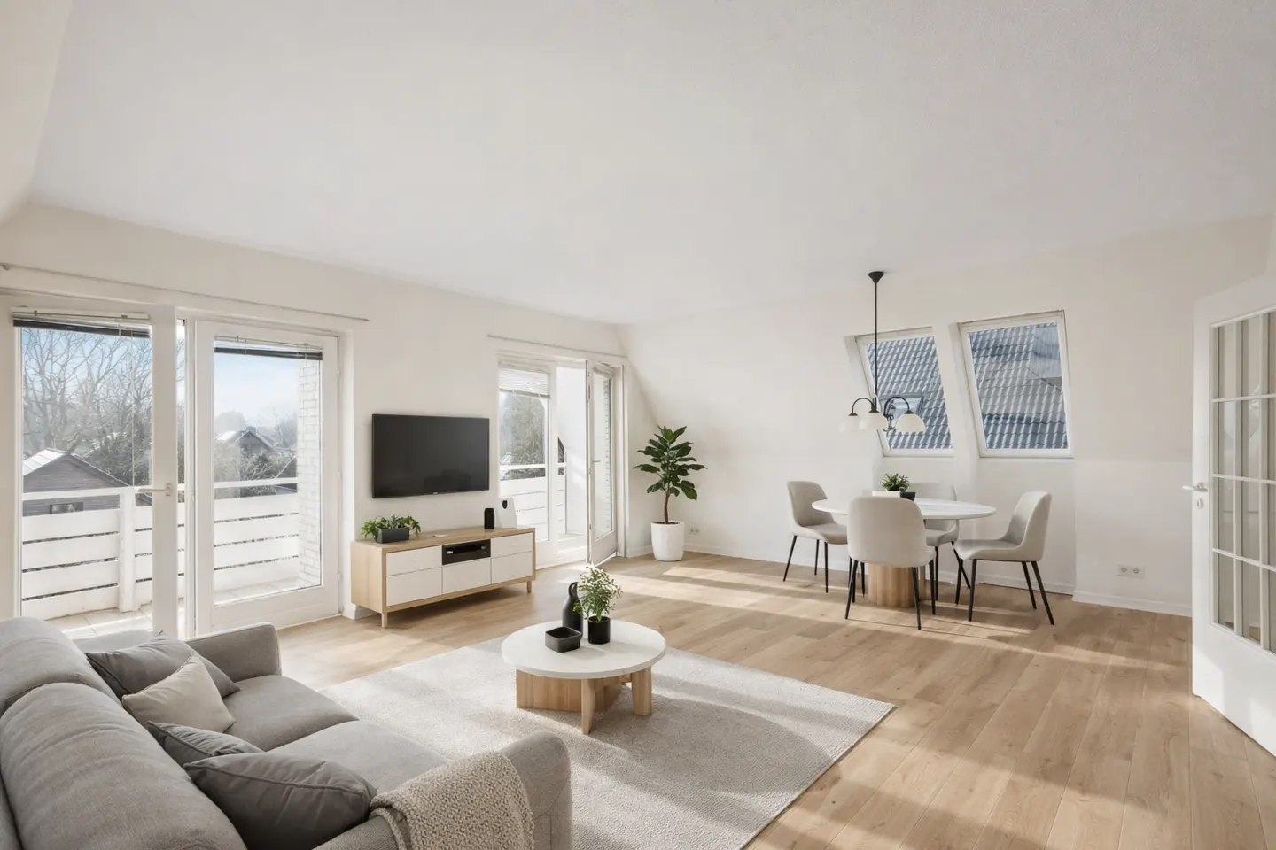 Bright, open living space with a gray sofa, round coffee table, TV, and dining set under a pendant light. Hardwood floors and white walls.