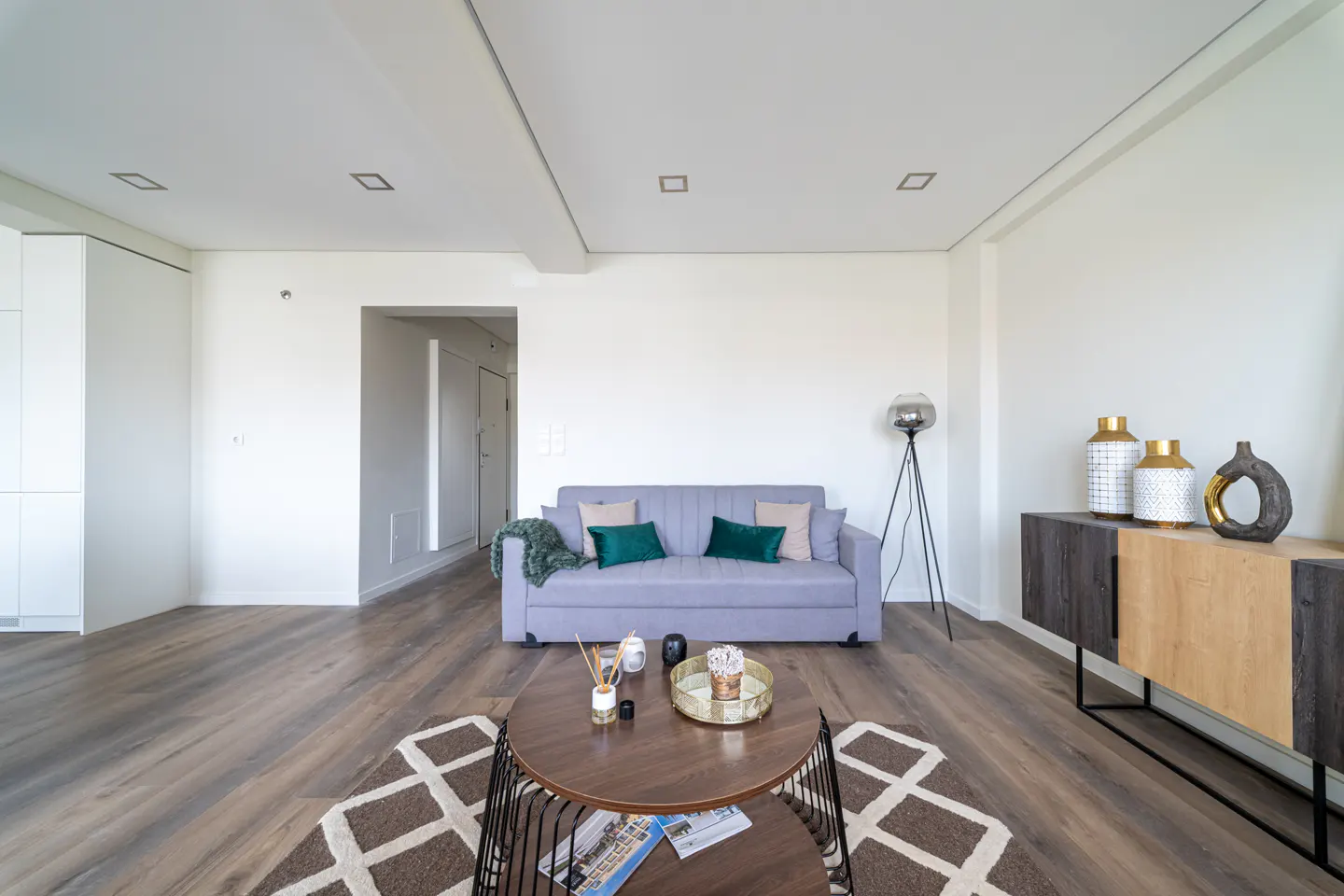 Living room with a gray sofa, green pillows, a round coffee table, and a patterned rug on a wood floor. Vases sit on a sideboard.