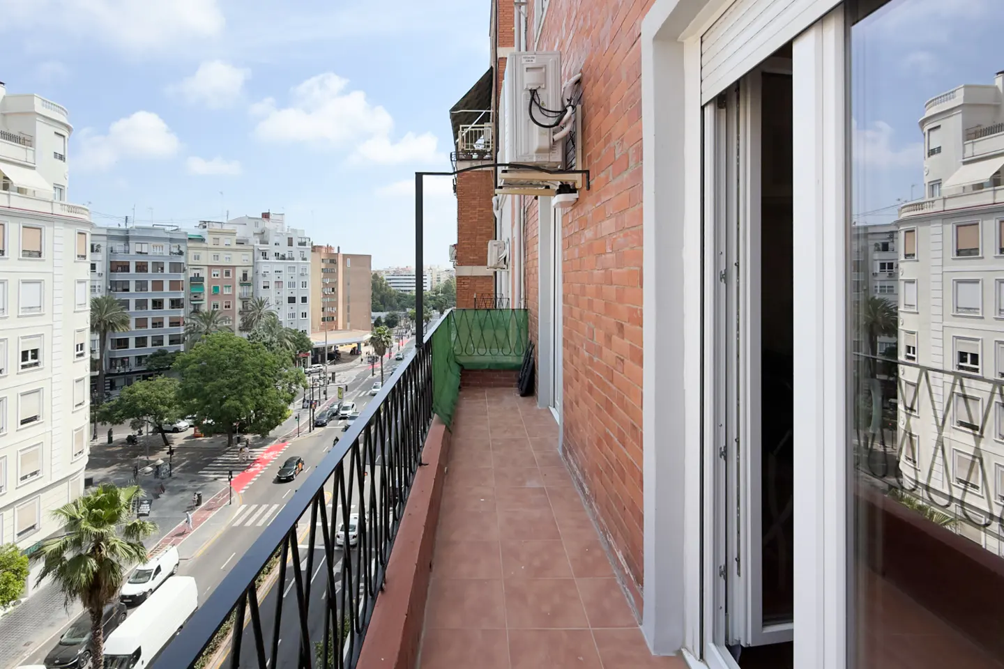 Balcony view of a city street. Black metal railing, terracotta tile floor, and brick wall. White sliding glass doors open to the city view.