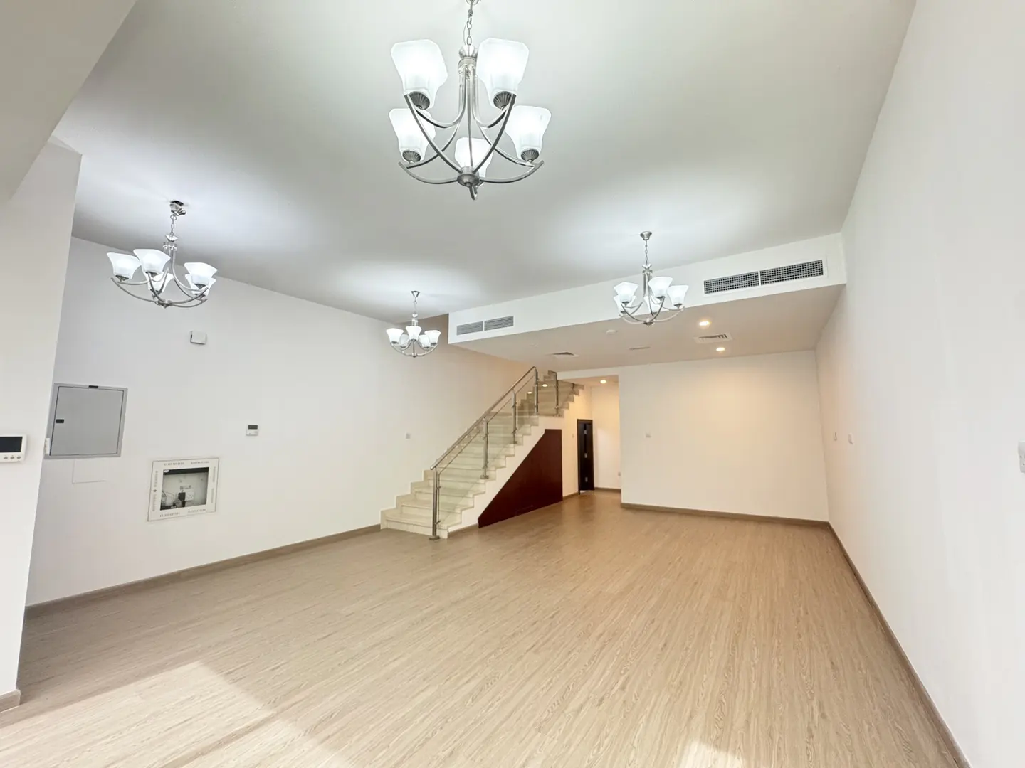 A bright, empty living room with light wood floors, white walls, and three chandeliers. A staircase leads to the upper level.