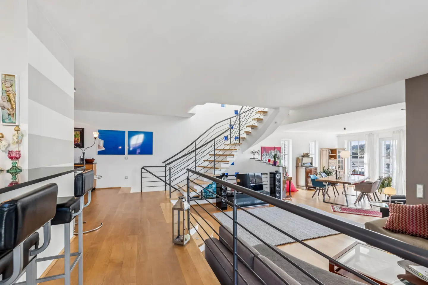Bright, modern living room with wood floors, white walls, and a curved staircase with black railings. Dining area and bar stools visible.