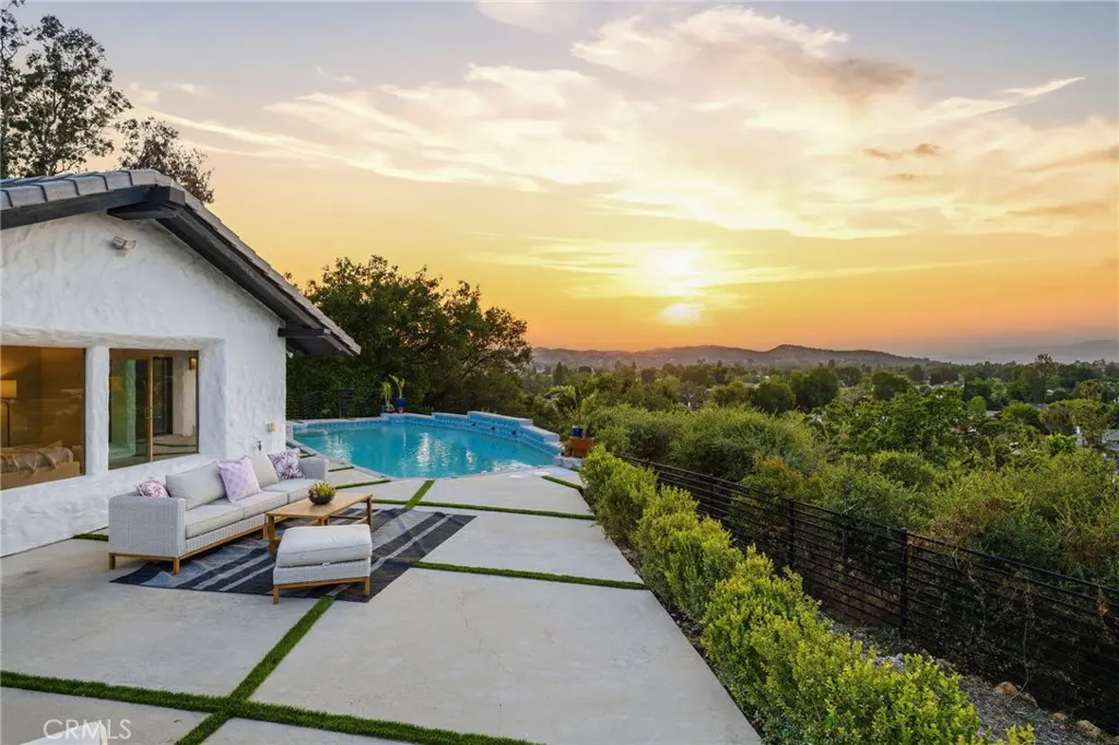 A white house with a pool and patio furniture overlooks a green landscape at sunset.