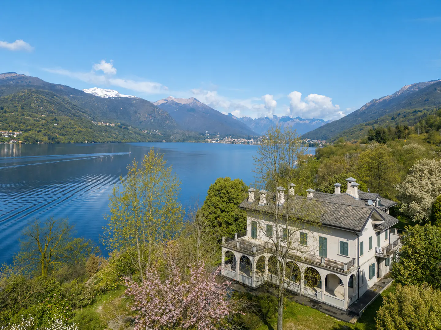 Aerial view of a large, light-colored house with a gray roof, overlooking a blue lake and green mountains under a clear blue sky.