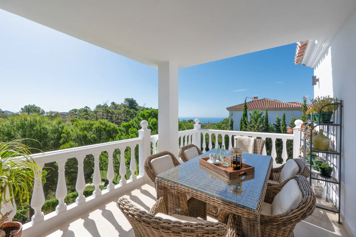 Outdoor patio with wicker table and chairs, white railing, and view of trees and blue sky.