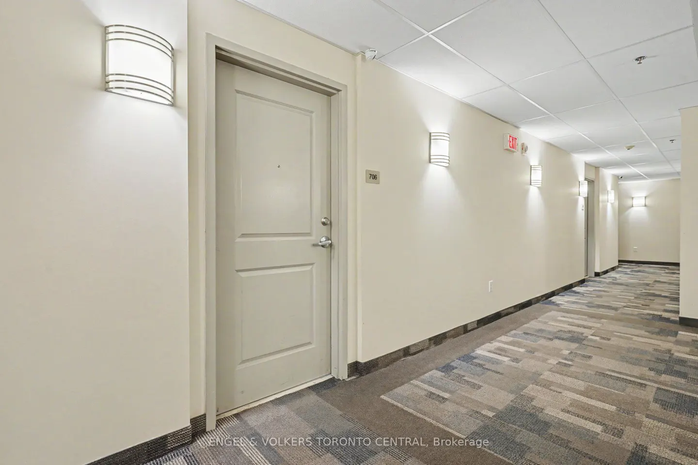 Hallway view with apartment door, number 706, and sconce lighting. Carpet is patterned with blue, gray, and beige. Cream colored walls.