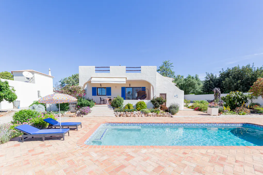 Exterior view of a white house with blue shutters, a pool, and lounge chairs on a sunny day.