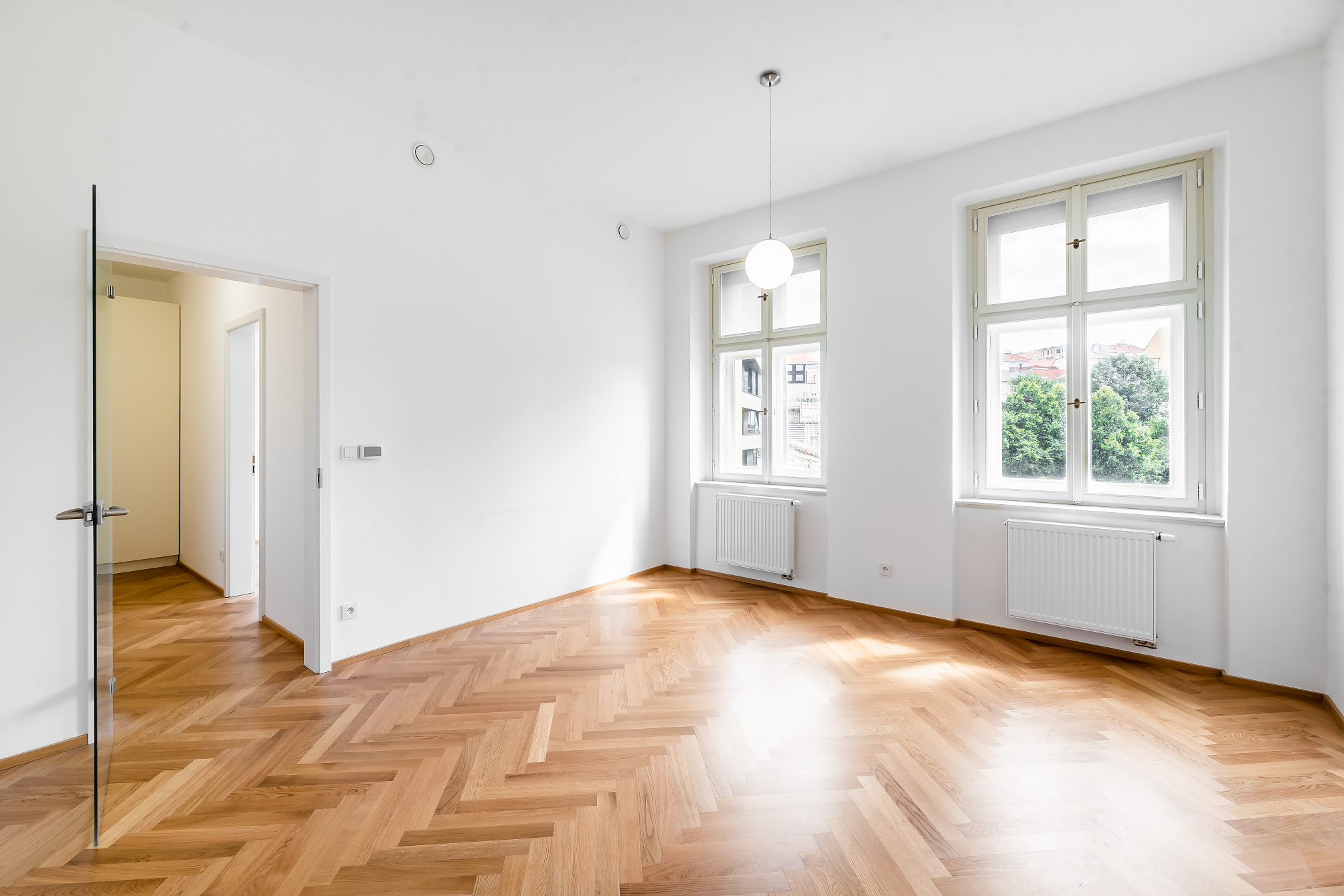 Empty room with white walls, herringbone wood floors, two windows with radiators, and a glass door leading to a hallway. [AI-generated text]