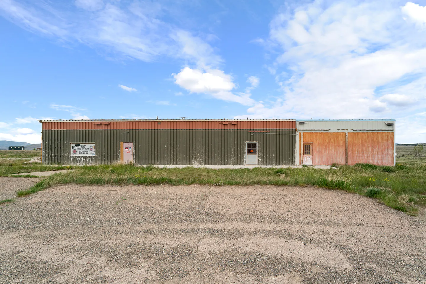 A long, single-story commercial building with olive and orange metal siding sits on a gravel lot under a blue sky.