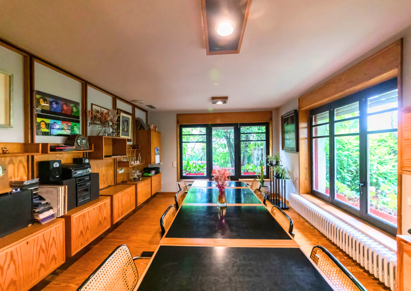 Bright room with a long black table, wood floors, and built-in wood shelving. Large windows look out to greenery.