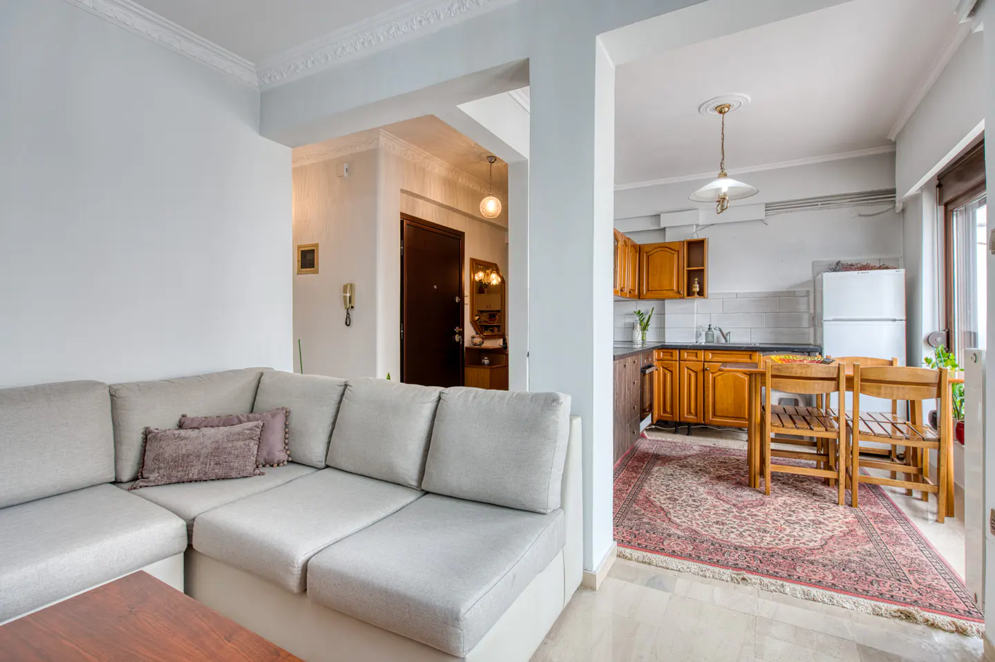 A bright apartment interior with a gray sectional sofa, a kitchen with wooden cabinets, and a dining table.