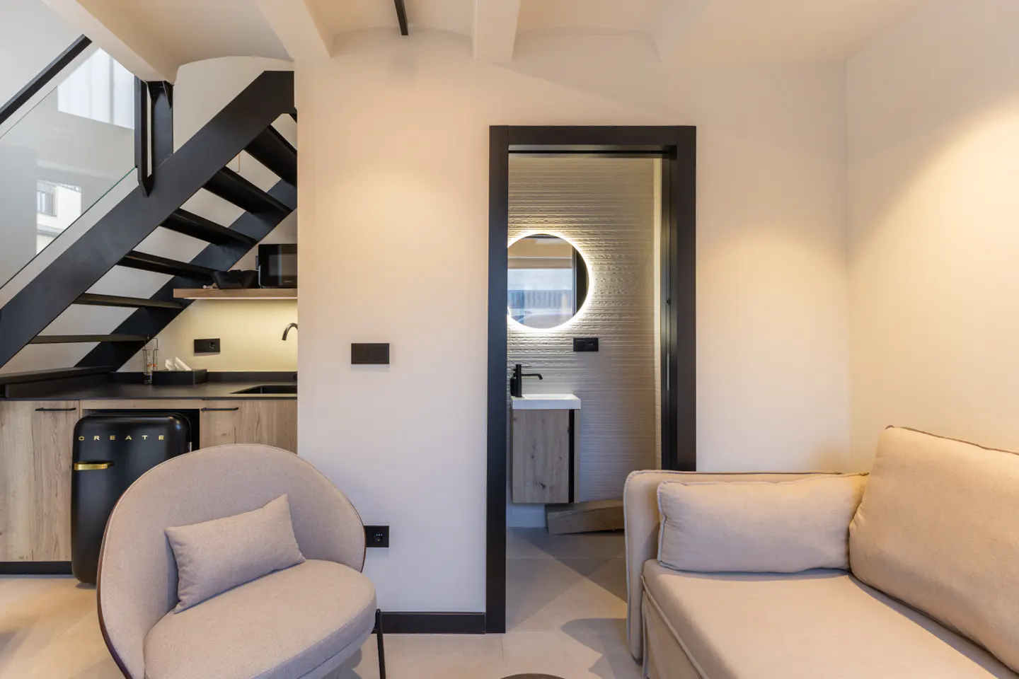 Interior view of a modern apartment with a black staircase, kitchenette, chair, sofa, and bathroom with a round mirror.
