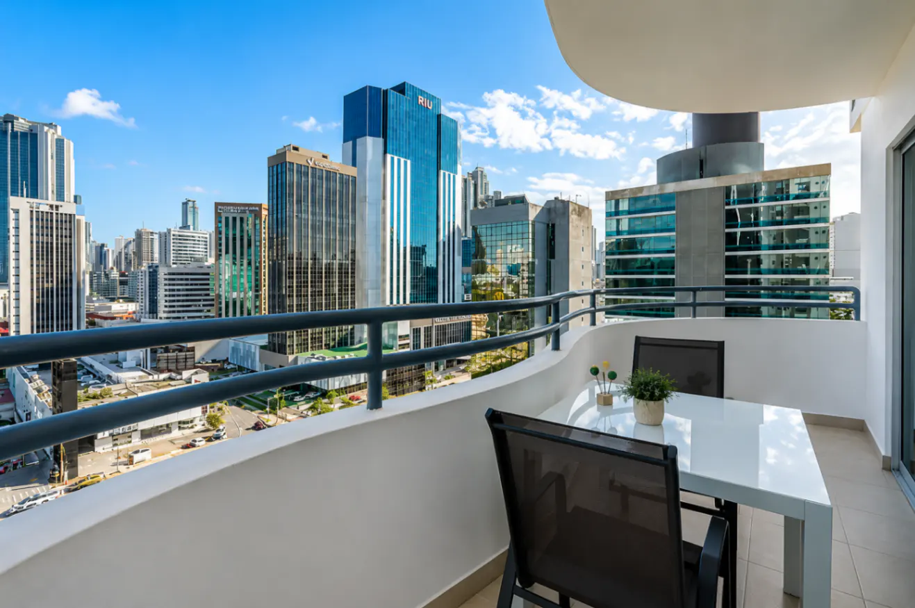 Balcony view of a city skyline. White table and two chairs sit on the balcony. Tall buildings are visible in the background.