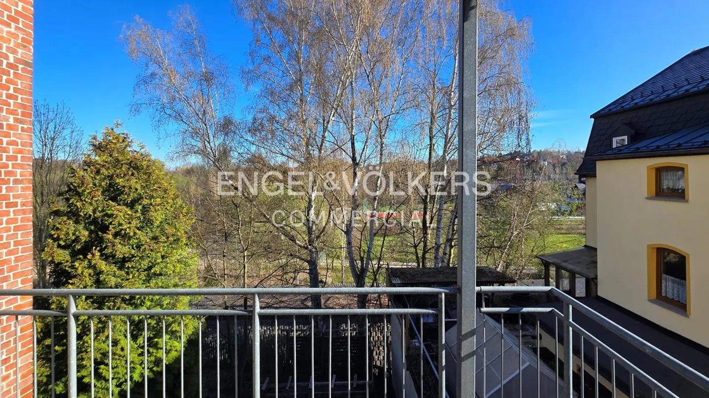 View from a balcony with metal railings, overlooking trees and a building with a black roof on a sunny day.