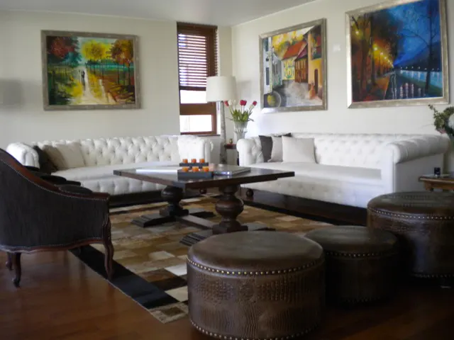 Living room with white sofas, brown leather ottomans, and paintings on the wall. A dark wood table sits on a patterned rug.