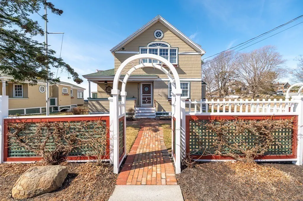Exterior view of a tan two-story house with a white arched gate and brick walkway. A white picket fence with red trim surrounds the property.