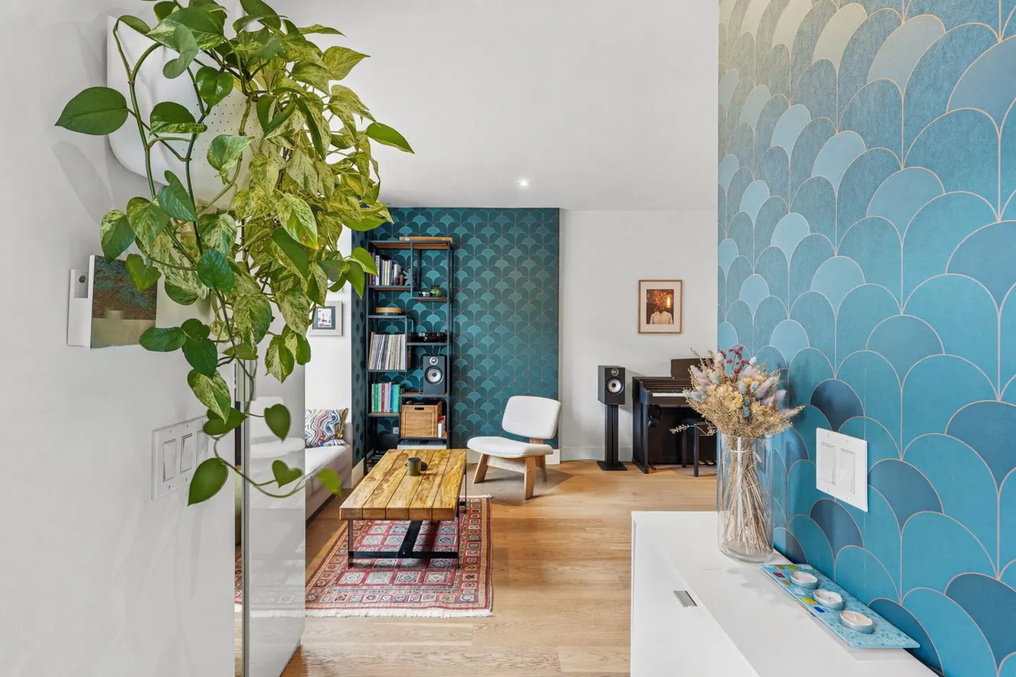 Living room with blue wallpaper, wood floors, and a hanging pothos plant. A bookshelf, chair, and piano are also visible.