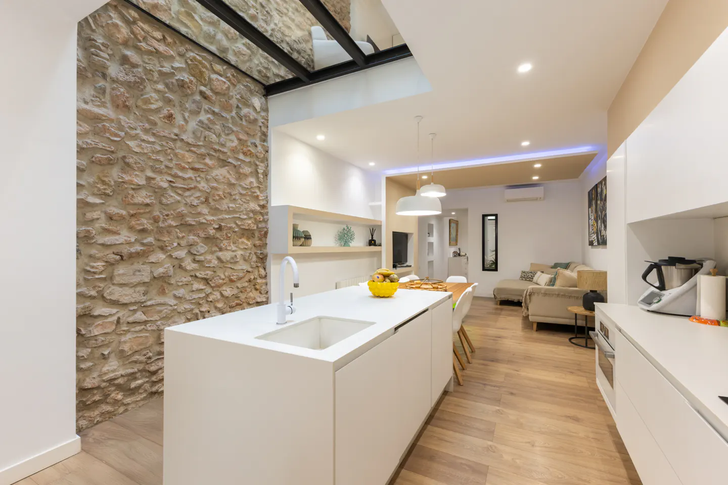 Bright, modern kitchen with white cabinets, island, and stone wall. Open floor plan leads to a living area with a beige sofa.