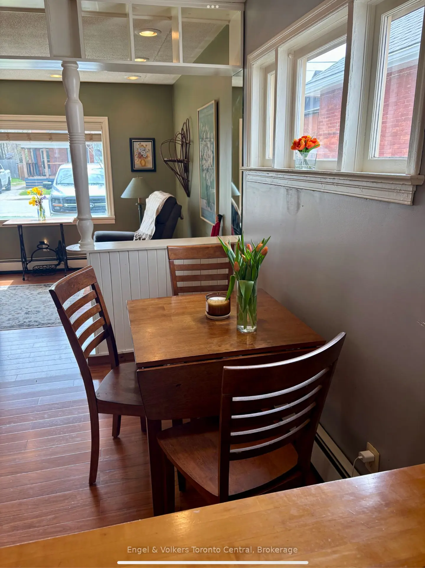 A dining area with a wooden table and chairs, featuring a vase of orange tulips. A window with a view of a residential street is visible in the background.
