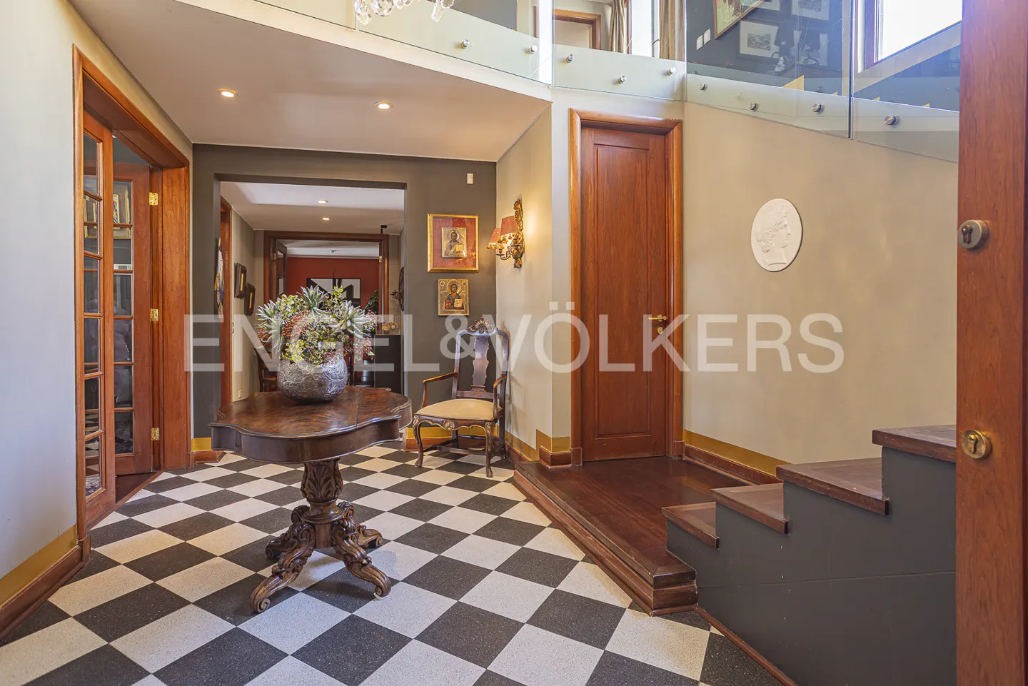 Elegant foyer with black and white checkered floors, a carved wooden pedestal table, and a staircase with glass railings.