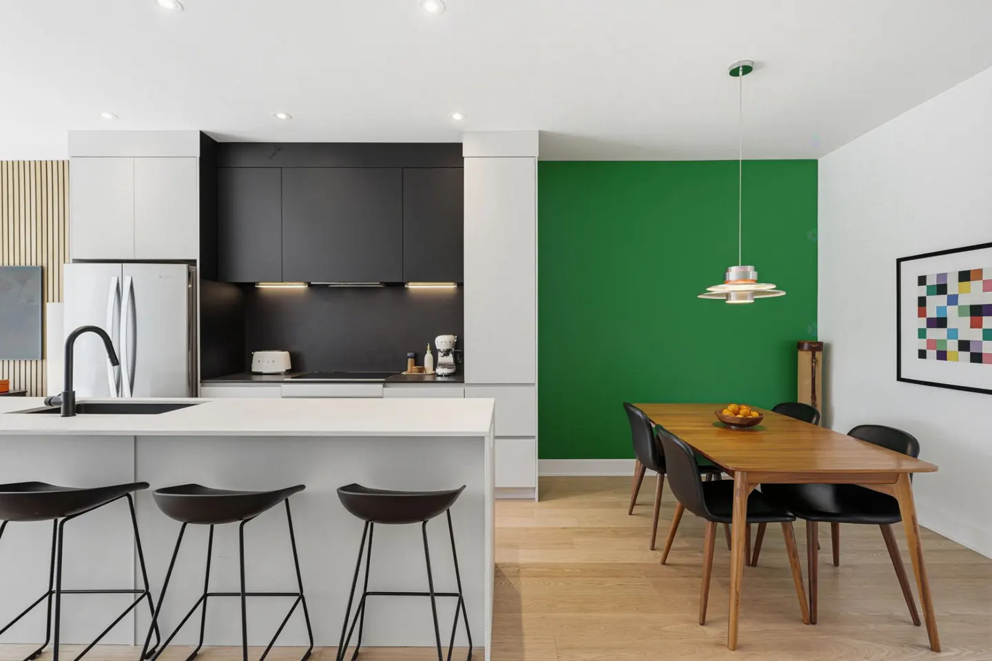 A modern kitchen with black and white cabinets, a white island with black stools, and a wooden dining table with black chairs against a green wall.