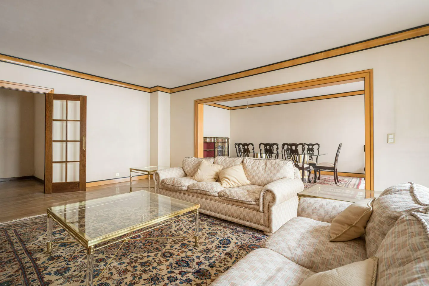 Living room with patterned sofas, glass coffee table, and a dining room visible through an opening. The walls are cream-colored.