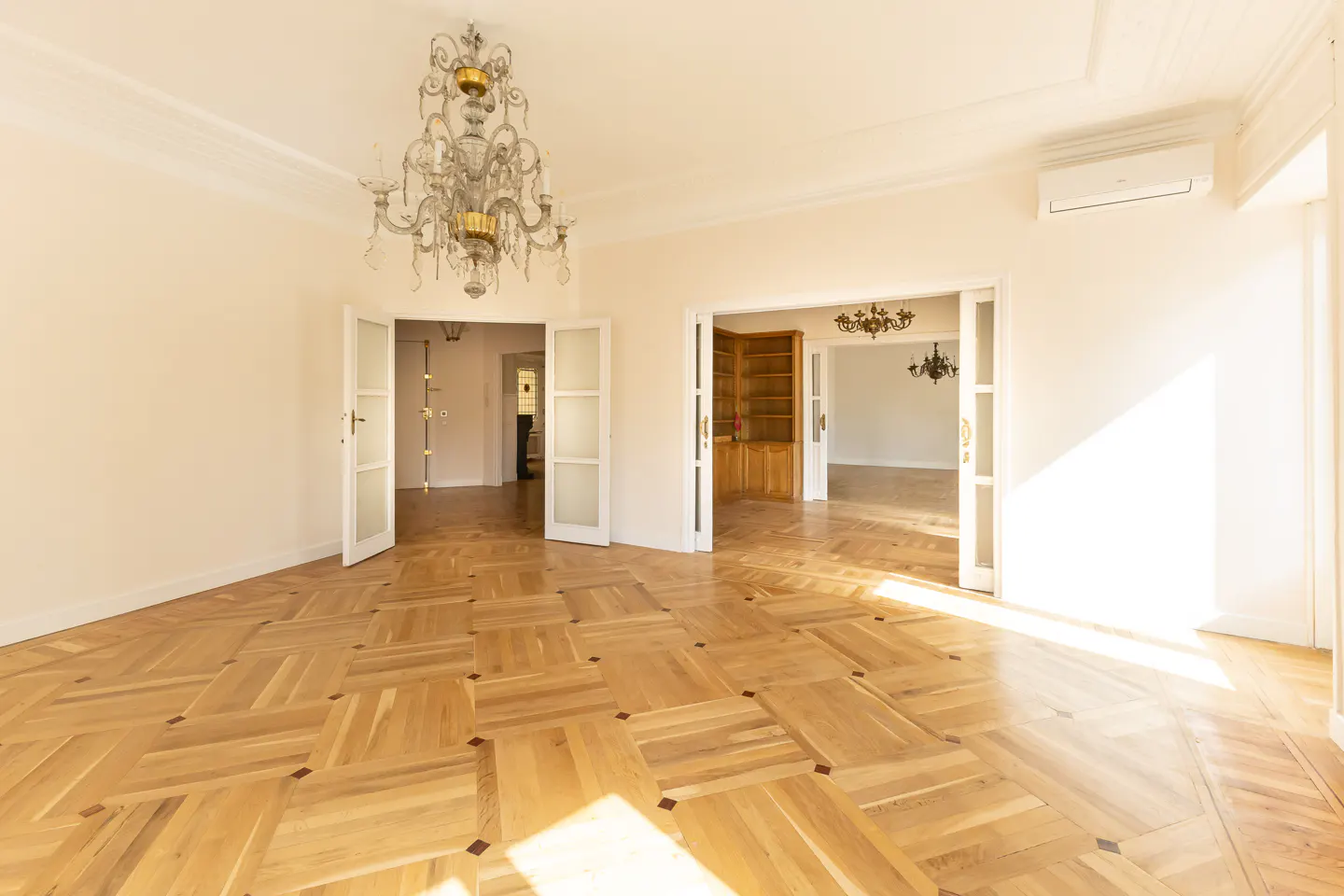 Bright, empty room with parquet floors, white walls, and open doorways. A chandelier hangs from the ceiling.