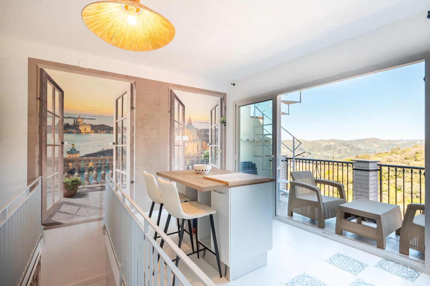 Bright room with a kitchen island, chairs, and a balcony with a view of rolling hills. A mural of a cityscape is on the wall.