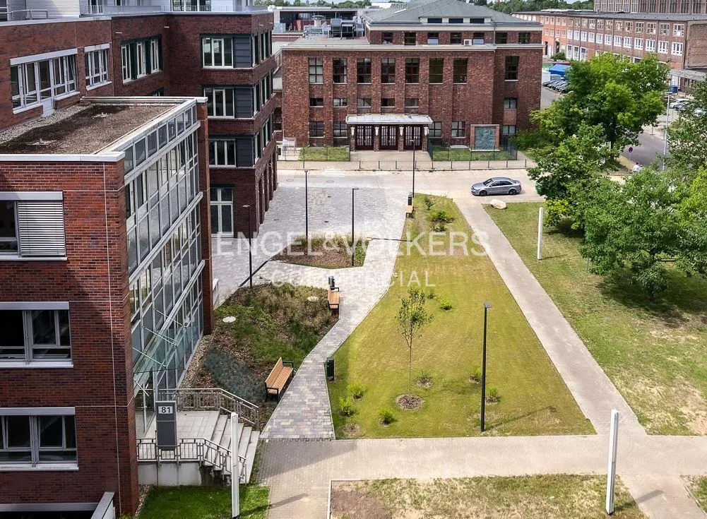 Aerial view of red brick buildings with green spaces and walkways. A silver car is parked near a building entrance. The Engel & Volkers logo is superimposed.