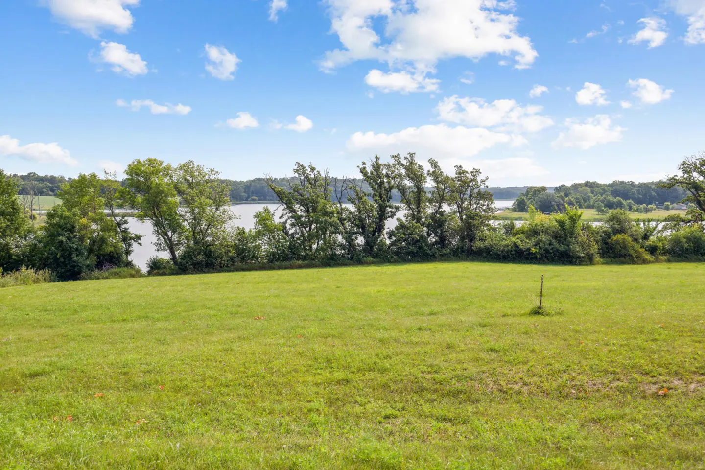 A green field with trees and a lake in the background under a blue sky with white clouds.