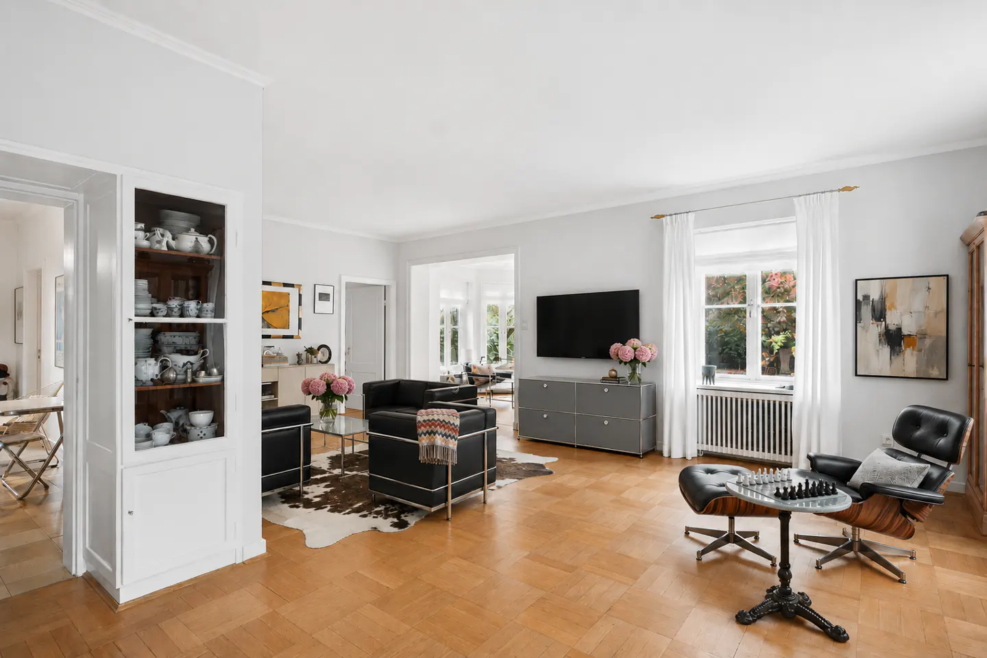 Bright living room with wood floors, white walls, and black leather chairs. A chess set sits next to a lounge chair. A cowhide rug adds texture.