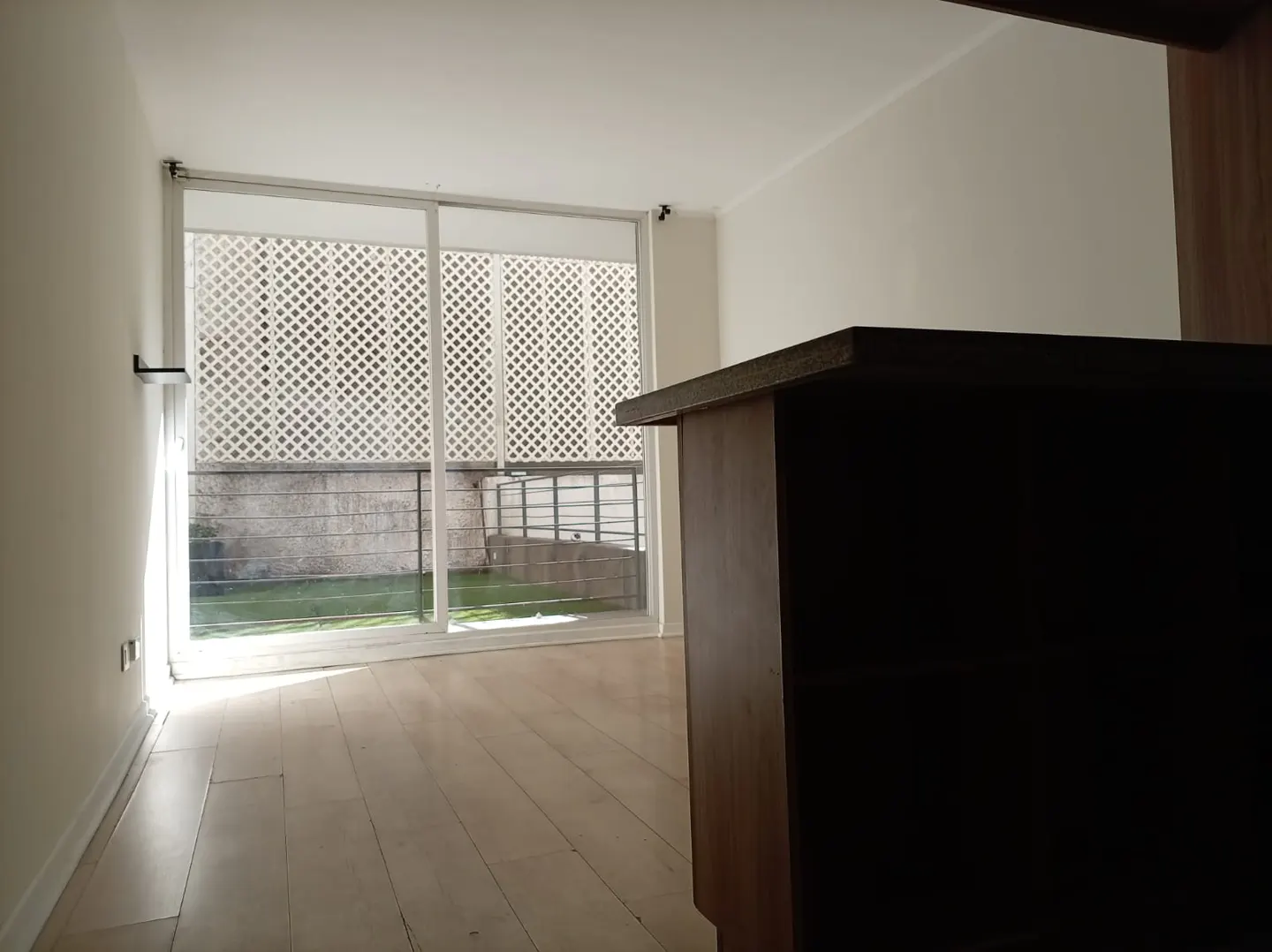 Bright, empty room with light wood floors and white walls. A large window with white lattice overlooks a green lawn. Dark wood cabinet in foreground.