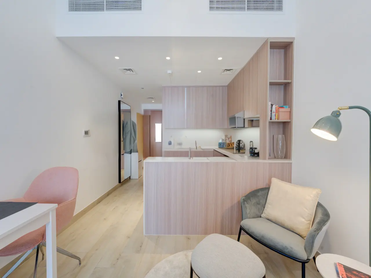 Bright, modern apartment interior with light wood cabinets, pink chairs, and a gray armchair under a green lamp.