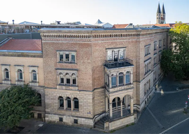 Exterior view of a tan brick building with arched windows and a small balcony, with church spires in the background.