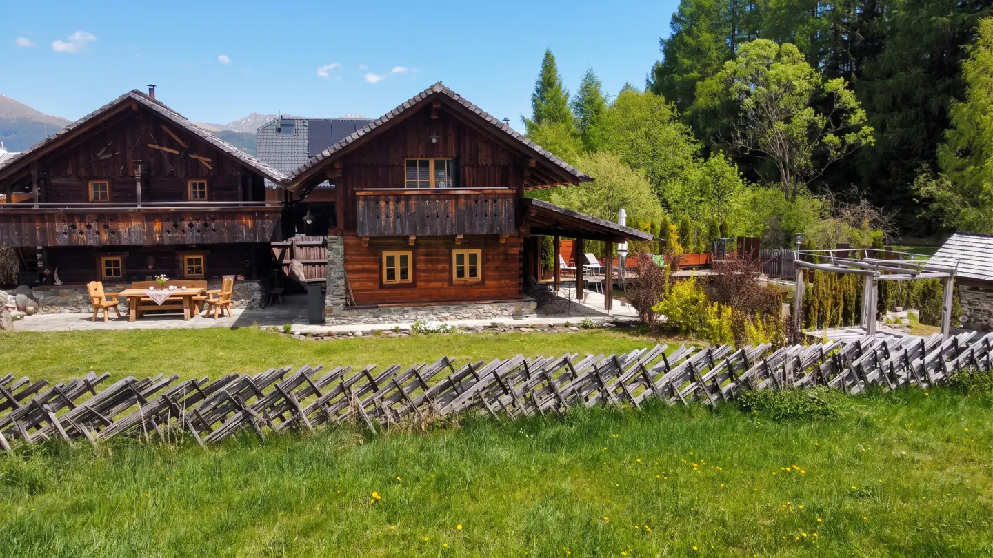Two-story brown log cabin with a wooden fence, green grass, and trees in the background.