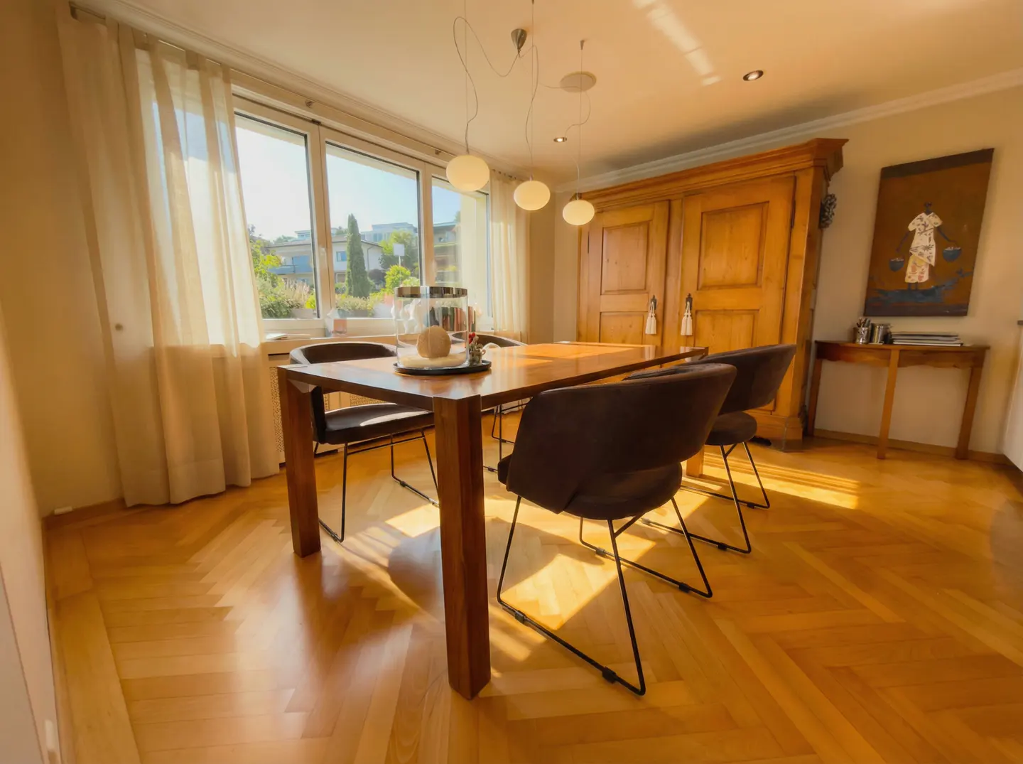 A dining room with a wooden table and four chairs, a large window with sheer curtains, and a wooden cabinet.