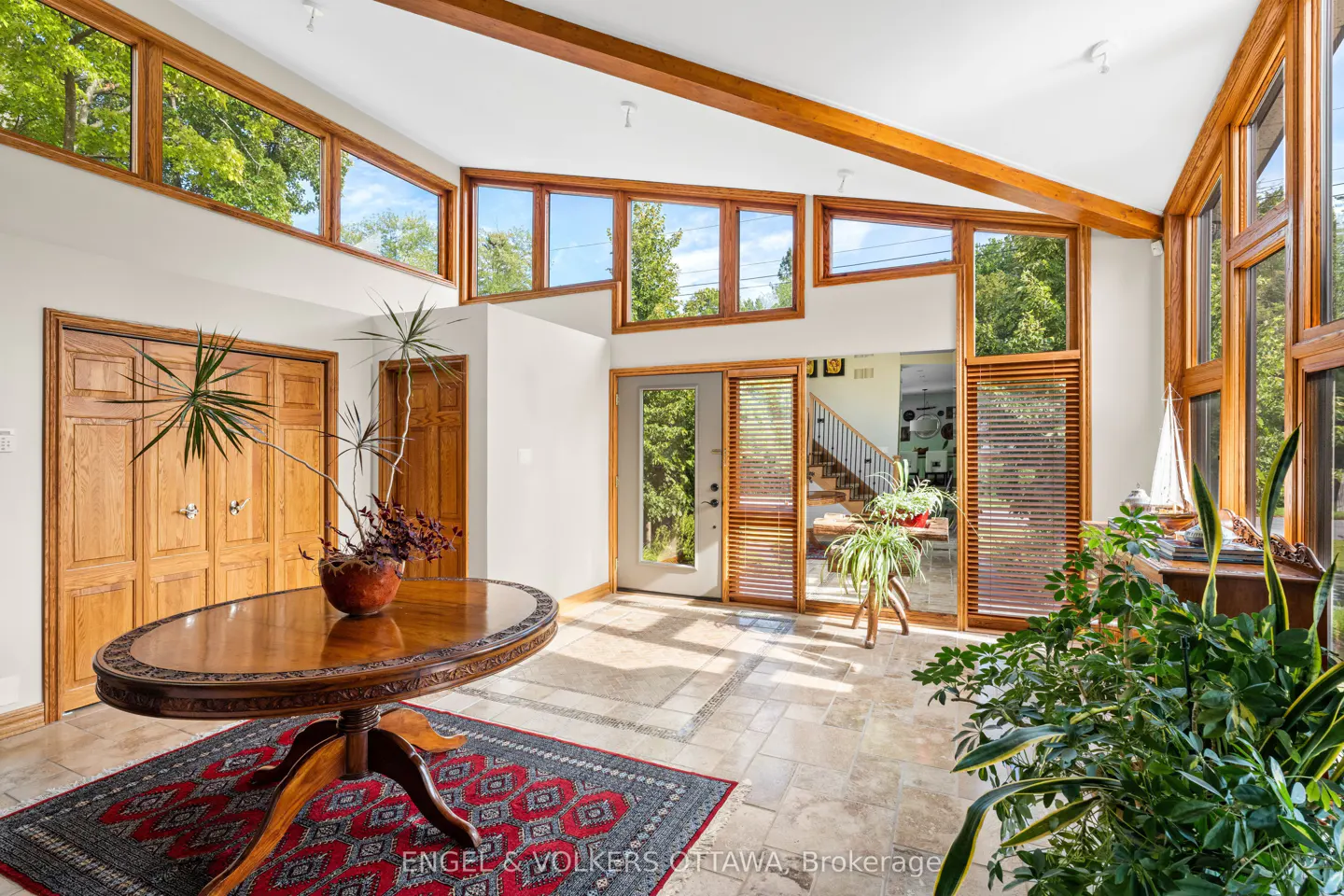 Bright foyer with wood beams, many windows, and a round table with a red rug. A staircase is visible in the background.