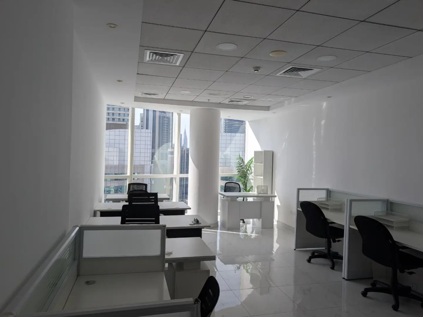 Bright, empty office space with white walls, desks, and black chairs. Large windows offer a city view.